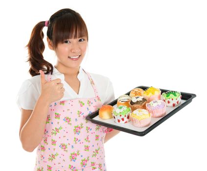 Thumb up Young Asian female baking bread and cupcakes, wearing apron and gloves holding tray isolated on white.