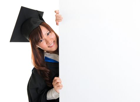 Portrait of smiling Asian female student in graduate gown peeking, hiding behind a white card, isolated on white background