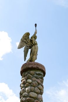 Angel holding torch on cloudy sky background. Statue on platform made of stones.