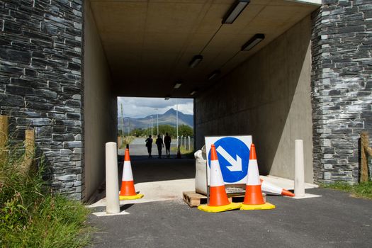 A closed single track road underpass with bollards and an arrow sign and three people walking through towards a mountain in the distance.