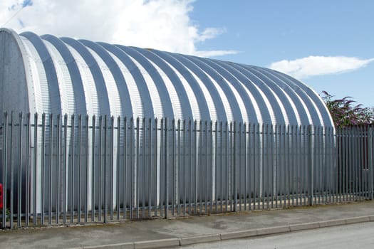 An industrial aluminium building behind security railings with a blue sky with cloud.