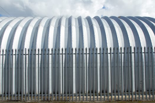 An industrial aluminium building behind security railings with a blue sky with cloud.