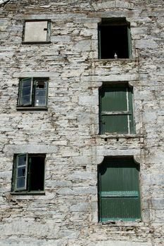 The frontage of a derelict, abandoned, building with broken windows and shutters and a white pigeon in an opening.