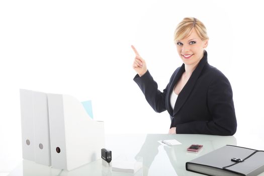 Smiling professional businesswoman seated at her white glass topped desk pointing to blank copy space above and to the side 