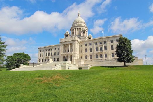 Front view of the Rhode Island state capitol building and lawn on Capitol Hill in Providence with bright blue sky and white clouds in the background. The building is covered with white Georgia marble and was constructed from 1895 to 1904.