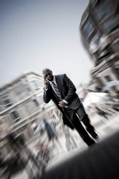 A confident business man walking through the city while talking on his smartphone. Zoom blur vignetting and desaturated toning for added effect.