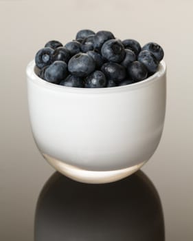 White glass cup full of blue blueberries reflecting on shiny gradient surface