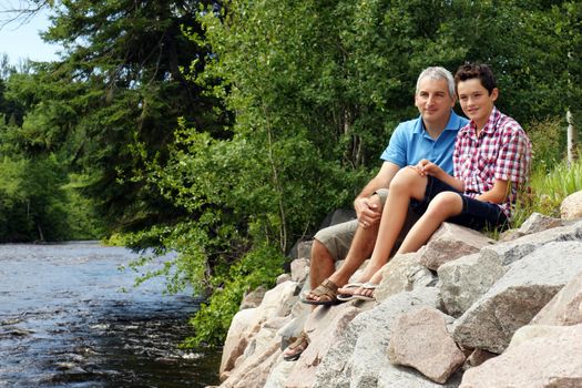 Beautiful shot of father and son sitting on rocks by the river enjoying summer at the cottage.