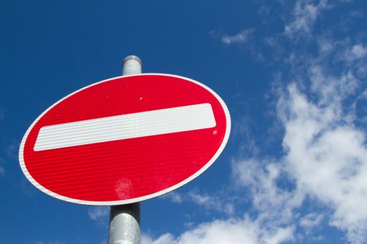 A no entry sign with a red circle and white bar on a metal post against a blue sky with cloud.