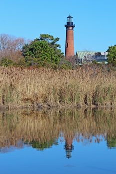 The red brick structure of the Currituck Beach Lighthouse near Corolla, North Carolina, is reflected in the calm waters of Currituck Sound against a bright blue sky