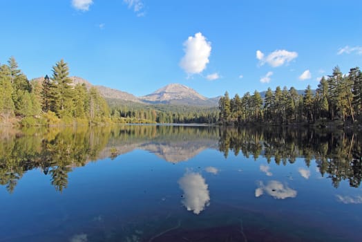 Wide-angle view of Lassen Peak, trees and white clouds reflected in the calm waters on Manzanita Lake near the entrance to Lassen Volcanic National Park in northern California