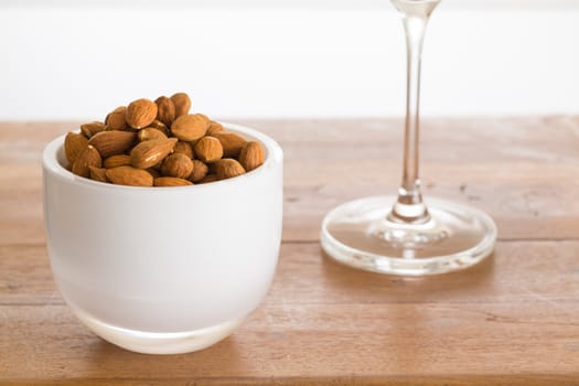 Raw almond nuts in white glass bowl on old wooden table with stem of wine glass