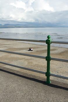 A set of railings over a sand and pebble beach with a couple sitting looking at clouds over the mountains in the distance.