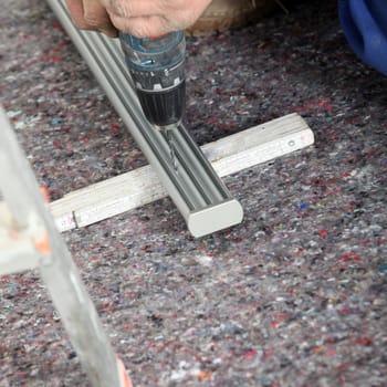 An artisan uses a portable cordless drill to drill a hole in a length of aluminium track supported at ground level.