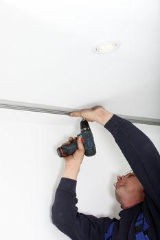 Man fitting a metal track to a white painted ceiling using a portable cordless drill