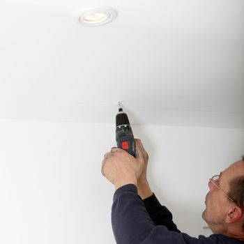 Man using a cordless battery-operated power drill to drill a hole in a white ceiling