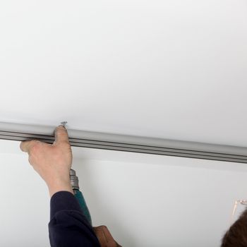 Man holding a length of metal track to a white painted ceiling while drilling a hole to attach it.