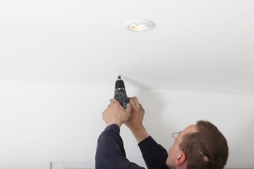 Man using a cordless battery-operated power drill to drill a hole in a white ceiling