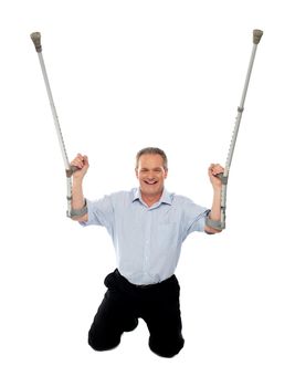 Cheerful senior man holding crutches upwards seated on floor, studio shot
