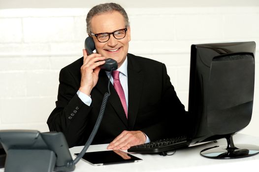 Senior businessman attending phone call at his work desk. Sitting in front of computer screen