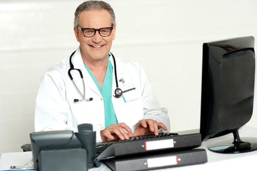 Aged male doctor in glasses working on computer, smiling at camera