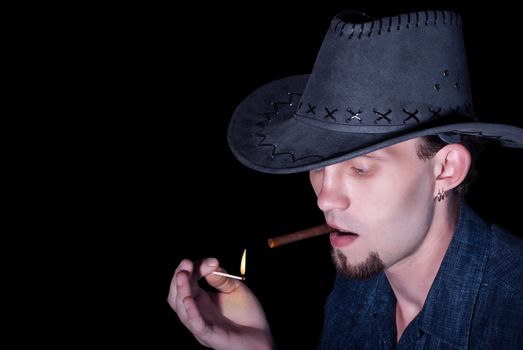 A young cowboy in a hat lights up a cigar on black background.