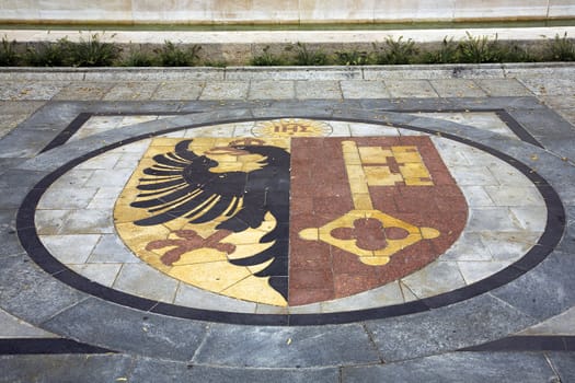 Coat of arms of the Swiss canton of Geneva embedded on the ground in front of the Reformation wall.  The black eagle is the emblem of Roman kingdom and the golden key is the symbol of Saint Peter the patron saint of the Cathedral of Geneva.  At the top a crest in the form of a sun within which are the letters IHS (or Σ, sigma), referring to Jhesus hominum salvator (Jesus, saviour of men).