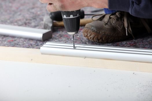 Drilling a hole using a handheld power drill in a piece of metal track on the ground.