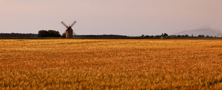 Evening landscape with a traditional windmill and modern irrigation tool in a field of cereals in the central region of France.