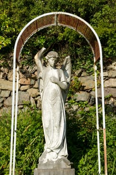 A stone carve statue of an angel, old and damaged under a metal frame with plants and a stone wall in the background.