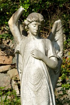 A stone carve statue of an angel, old, dirty and damaged with plants and a stone wall in the background.