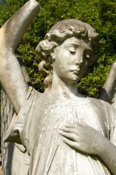 A stone carve statue of an angel, old, dirty and damaged with plants in the background.