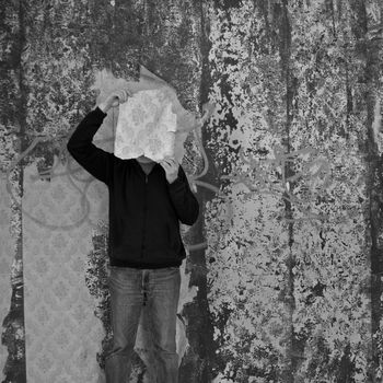 Man with piece of torn wallpaper in decayed room. Black and white.