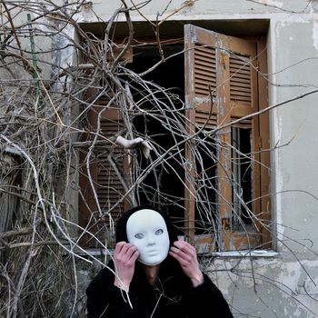 Grotesque white mask figure and haunted house window with tangled overgrown plants and creepy dummy doll hand.