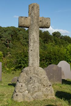 A stone carved cross and base in a graveyard with trees and a blue sky.