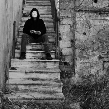 Masked figure with doll hand sitting on abandoned house staircase. Black and white.