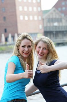 Happy female friends standing alonside an urban waterway giving an enthusiastic thumbs up of approval for their lifestyle Happy female friends standing alonside an urban waterway giving a thumbs up of approval for their lifestyle 