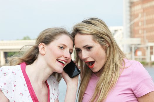 Two ladies standing with their heads close together listening to a conversation over a mobile phone, with one woman reacting in disbelief and shock 