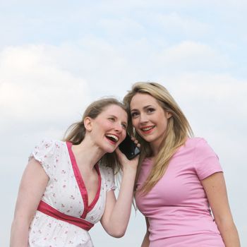 Two attractive woman standing with their heads close together listening to a phone call on the same mobile phone Two attractive woman standing with their heads close together listening to a phone call on the same mobile phone