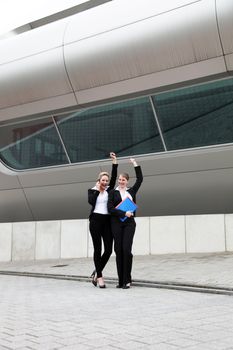 Two professional businesswoman in formal slacksuts standing in front of a modern corporate building waving 