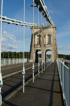 A section of a link chain leading to a stone tower with cables leading to a road as part of a suspension bridge with road and walkway.