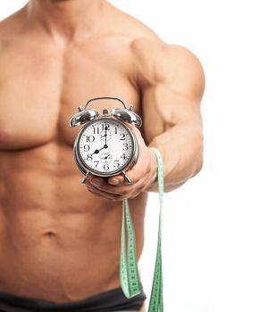 Cropped view of a muscular young man holding clock and measuring tape over white background. It is high time for workout concept.