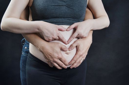 New daddy and mummy hands forming love shape on pregnant belly, over black background