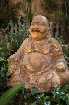 Terracotta Buddha Statue of a seated smiling Buddha with a jovial expression in greenery at the foot of a tree