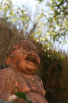 Low angle view of the face of a terracotta smiling Buddha statue outdoors looking up through greenery to blue sky