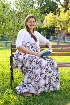 Beautiful woman pregnant sitting on a bench in a garden reading book