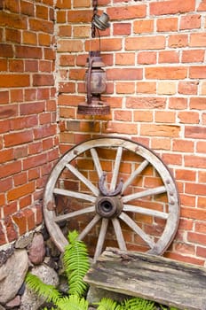 Antique wooden carriage wheel with binding of metal and horseshoe on it. Kerosine paraffin rusty lamp. Red brick wall. Fern leaves.