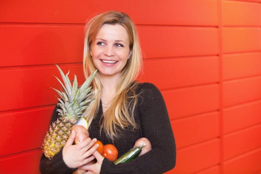 Smiling woman leaning against red wall while holing fruits and vegetables