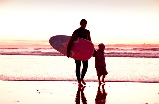 Female surfer and her daughter walking in the beach at the sunset