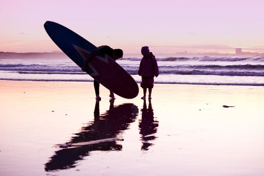 Female surfer and her daughter walking in the beach at the sunset